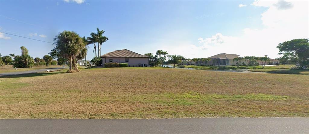 A vacant, grassy residential lot sits in front of a lake and neighboring houses — 17247 CARROWAY LANE, PUNTA GORDA, FL