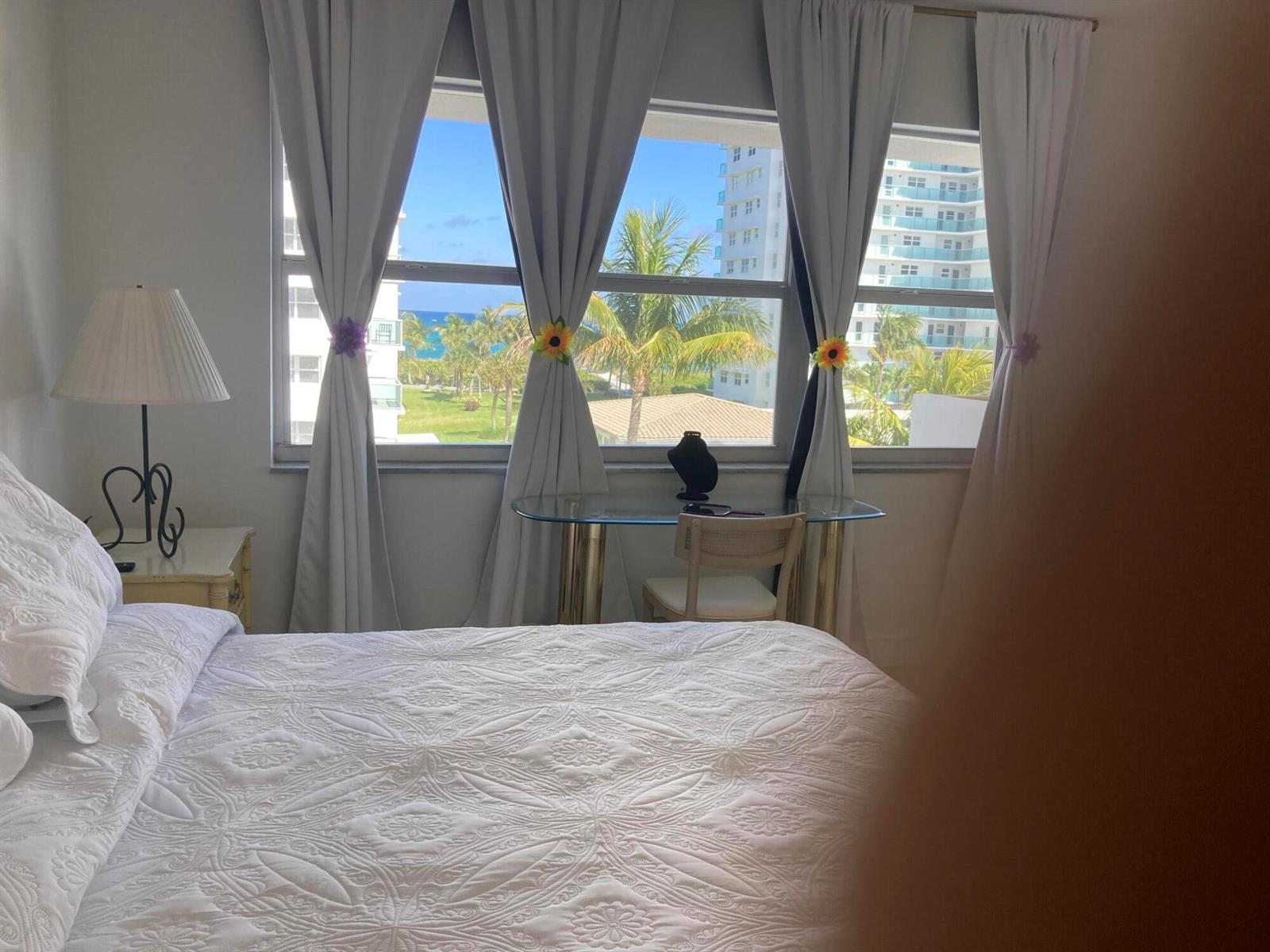 A bedroom features a white quilt, a glass desk, and a window view of the ocean — 1630 N Ocean Boulevard # 314, Pompano Beach FL 33062, POMPANO BEACH, FL