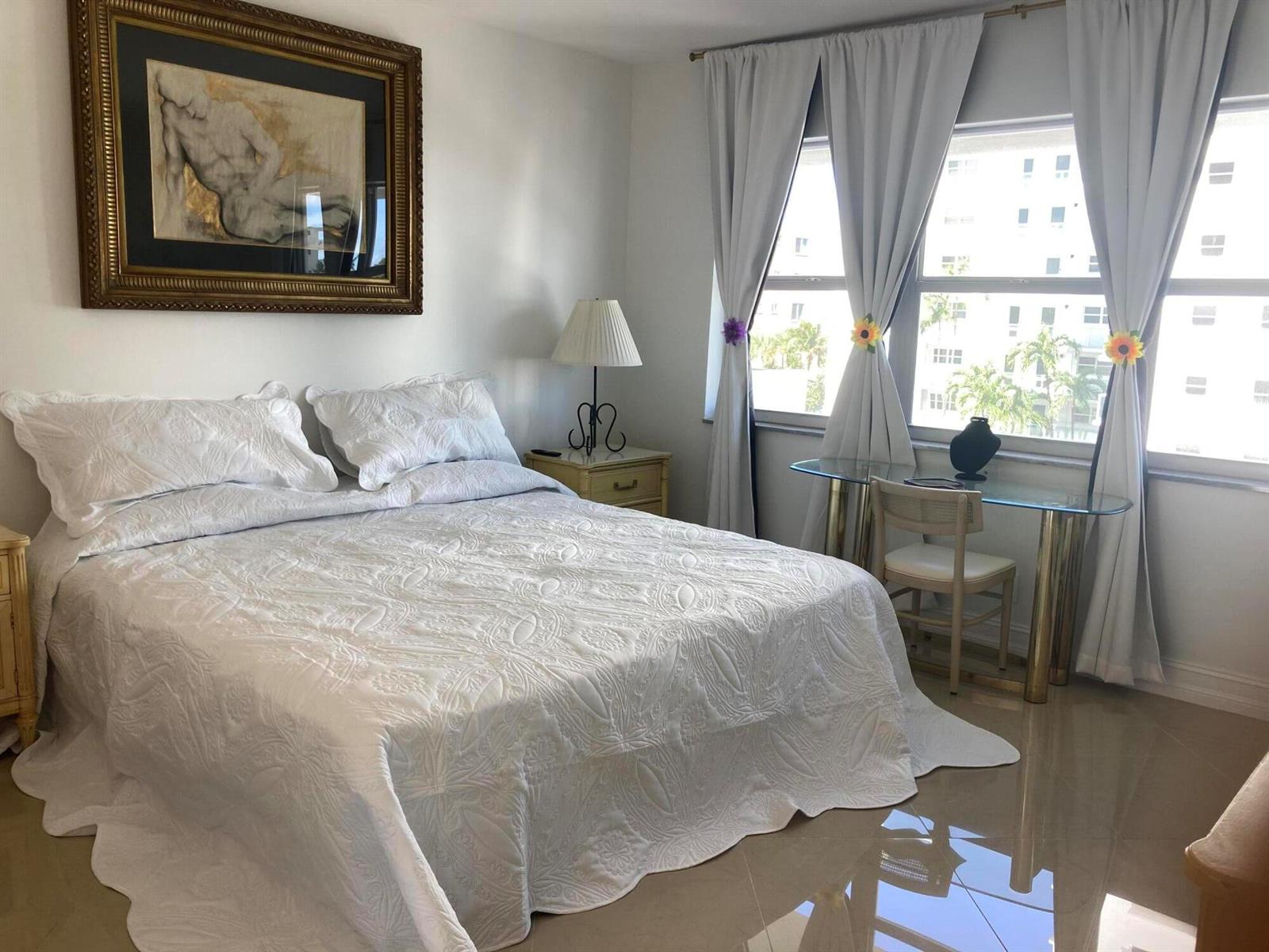 A bedroom features a white quilted bed, a glass desk, and a large framed painting — 1630 N Ocean Boulevard # 314, Pompano Beach FL 33062, POMPANO BEACH, FL