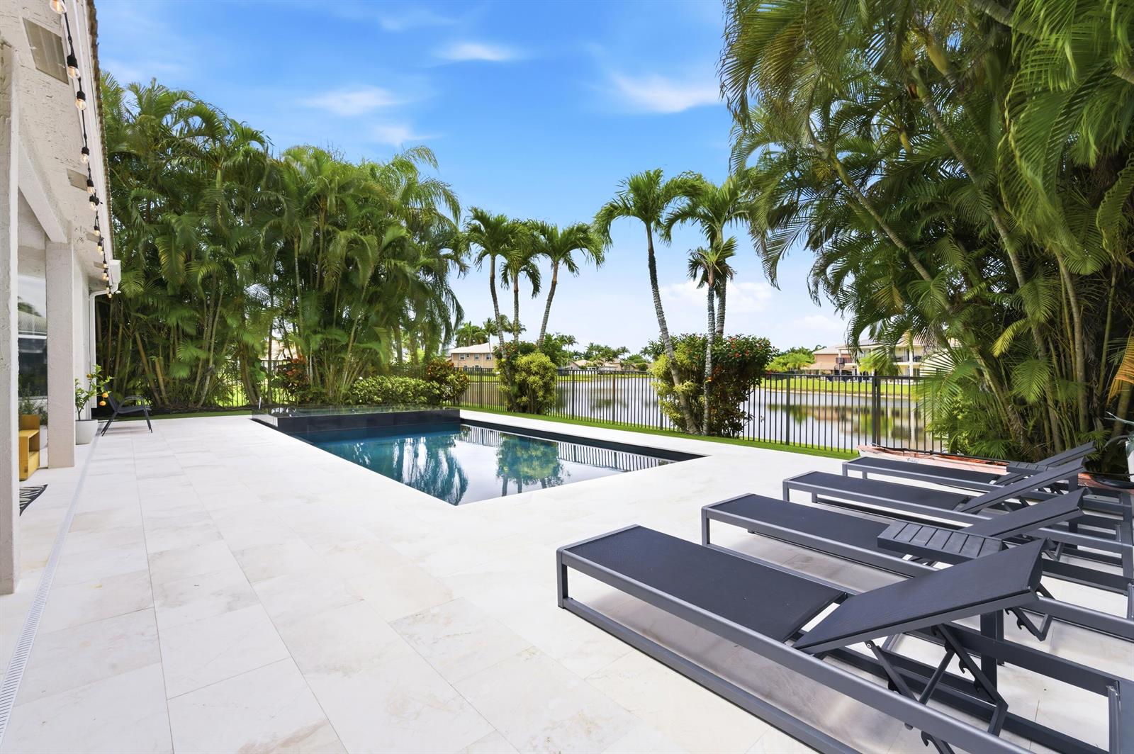 A backyard patio features a rectangular swimming pool, lounge chairs, and palm trees by a lake — 19123 Two River Lane, Boca Raton FL 33498, BOCA RATON, FL