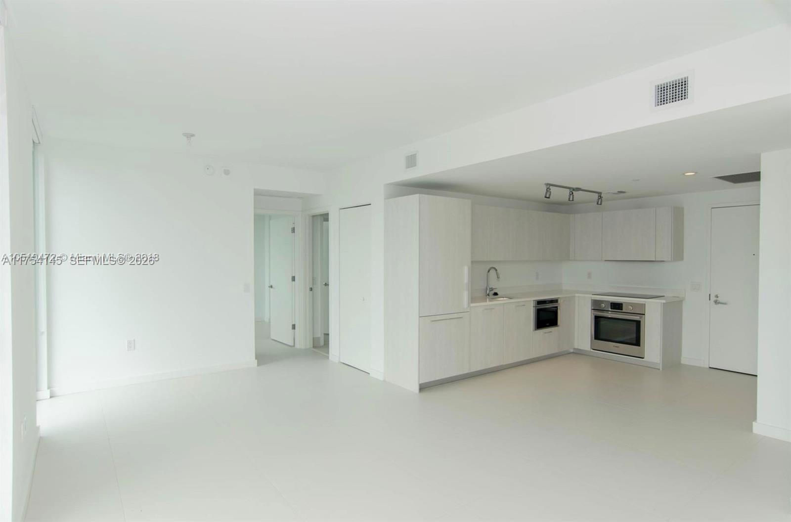 A modern, minimalist kitchen with white cabinetry and stainless steel appliances opens into a living area — 501 NE 31st St # 2009, Miami FL 33137, MIAMI, FL