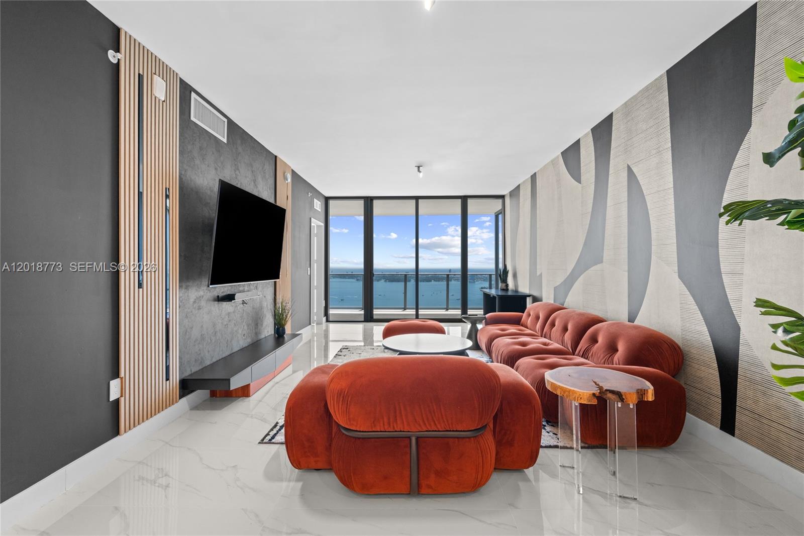 A modern living room features a rust-colored sofa, a wall-mounted television, and floor-to-ceiling windows — 700 NE 24th St # 5206, Miami FL 33137, MIAMI, FL