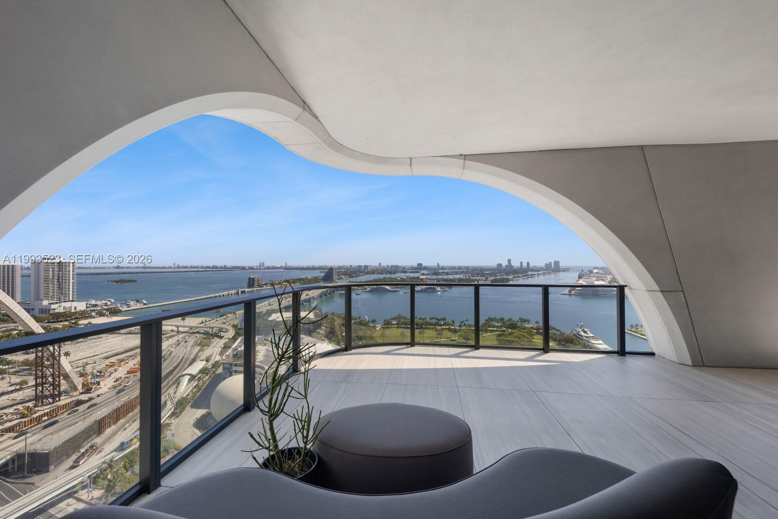 A high-rise balcony features a curved architectural ceiling, glass railings, and panoramic waterfront views — 1000 Biscayne Blvd # 2702, Miami FL 33132, MIAMI, FL