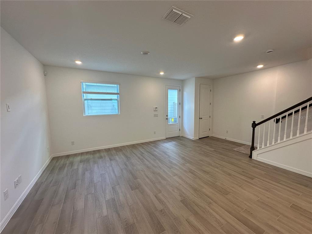 An empty living room features light wood-look flooring, white walls, a staircase, and a door — 6541 CYRILS DRIVE, ST CLOUD, FL