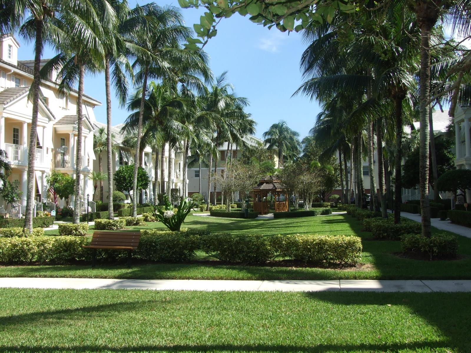 A grassy courtyard with palm trees, a wooden gazebo, and residential buildings under a blue sky — 1554 Limetree Bay Avenue, Jupiter FL 33458, JUPITER, FL