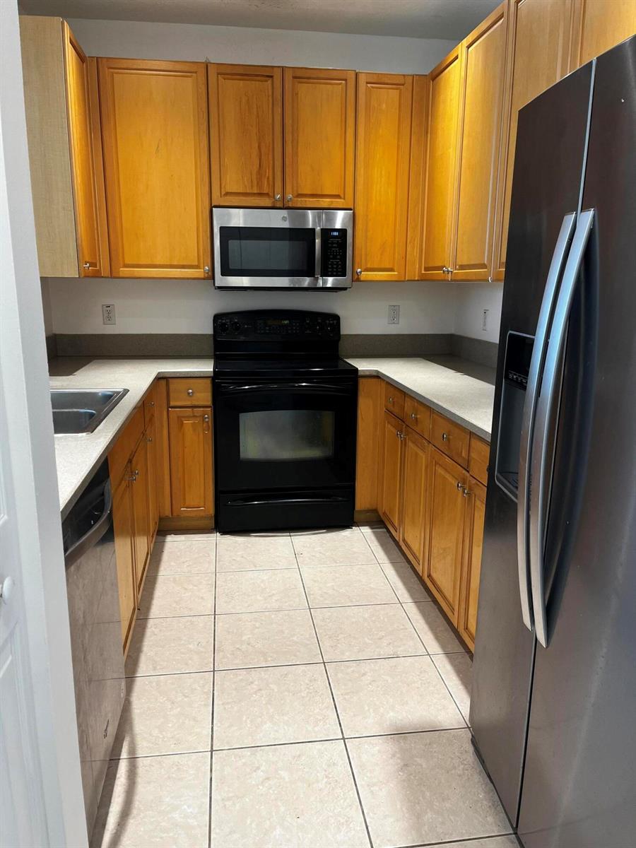 A galley kitchen features honey-oak cabinets, black appliances, and light-colored tile flooring — 1554 Limetree Bay Avenue, Jupiter FL 33458, JUPITER, FL