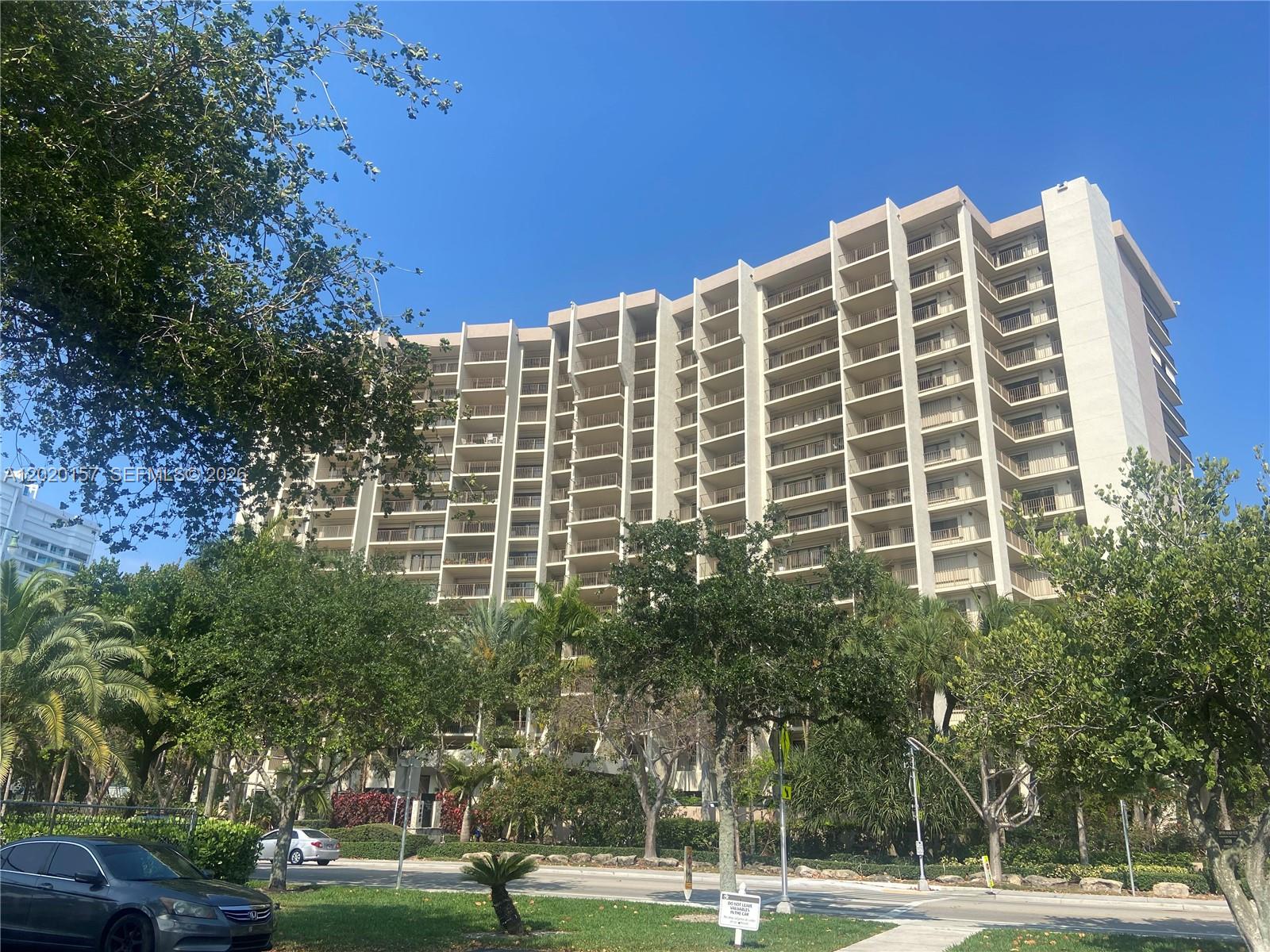 A tall, multi-story residential building stands behind lush green trees and a paved road — LAUDERDALE BY THE SEA, FL