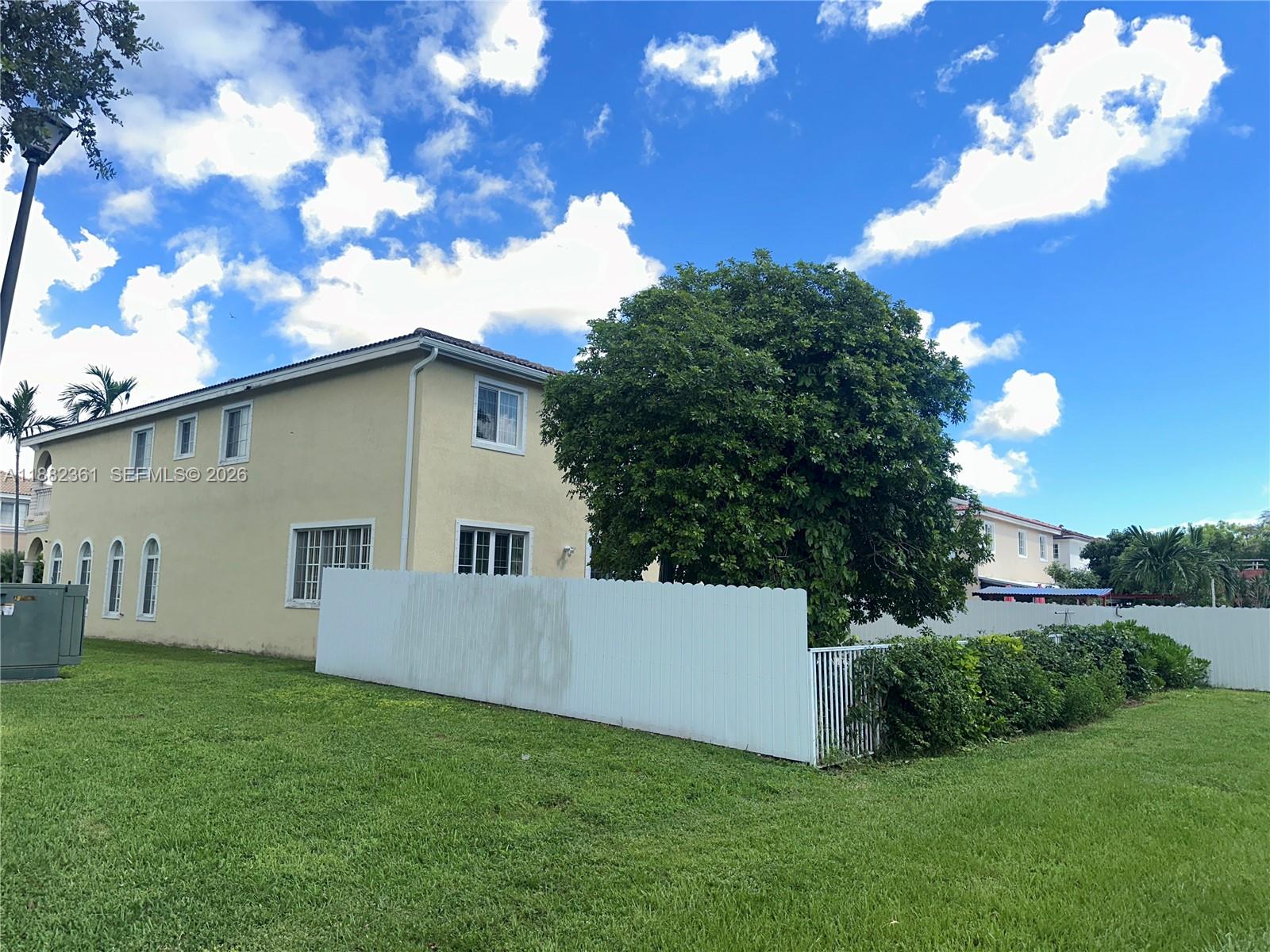 A two-story yellow house stands behind a white fence next to a large green tree — 13986 SW 276th Way, Homestead FL 33032, HOMESTEAD, FL