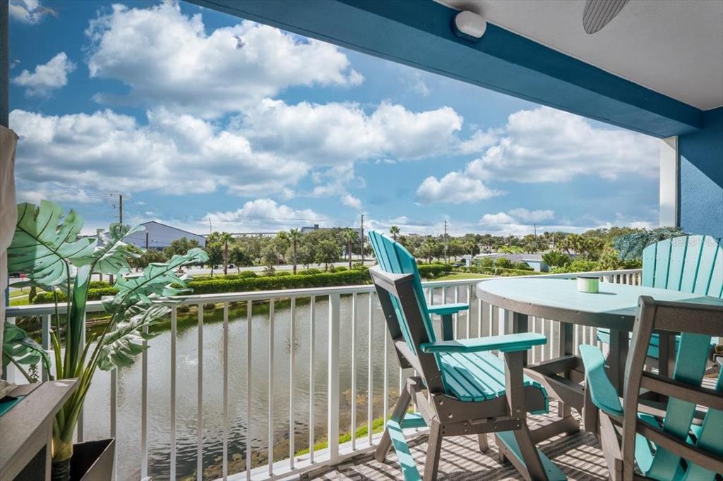A balcony features teal patio furniture overlooking a small pond and a cloudy blue sky — 1 RIVERWALK DRIVE UNIT 303, NEW SMYRNA BEACH, FL