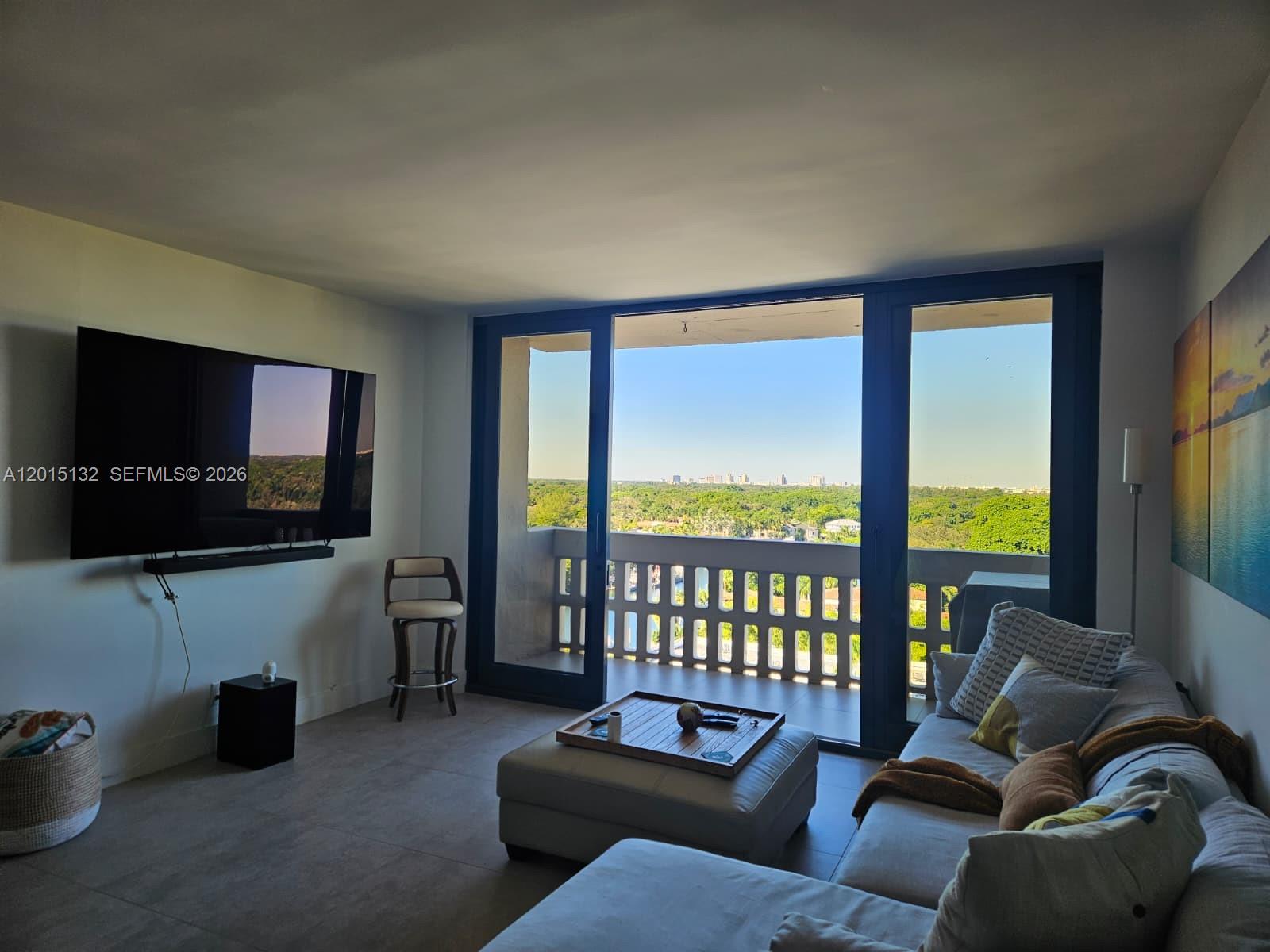 A living room features a wall-mounted television, a sectional sofa, and a balcony with views — 90 Edgewater Dr # 1219, Coral Gables FL 33133, CORAL GABLES, FL
