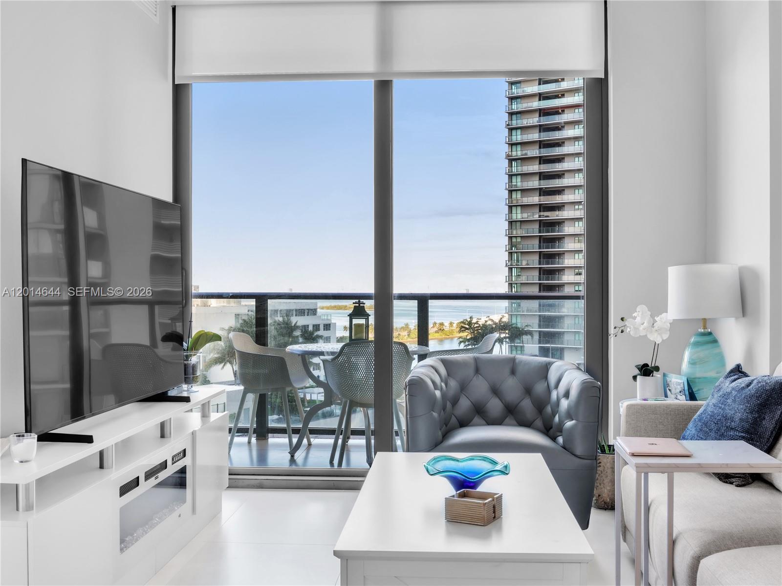 A modern living room features a white media console, gray tufted armchair, and balcony view — 501 NE 31st St # 1105, Miami FL 33137, MIAMI, FL