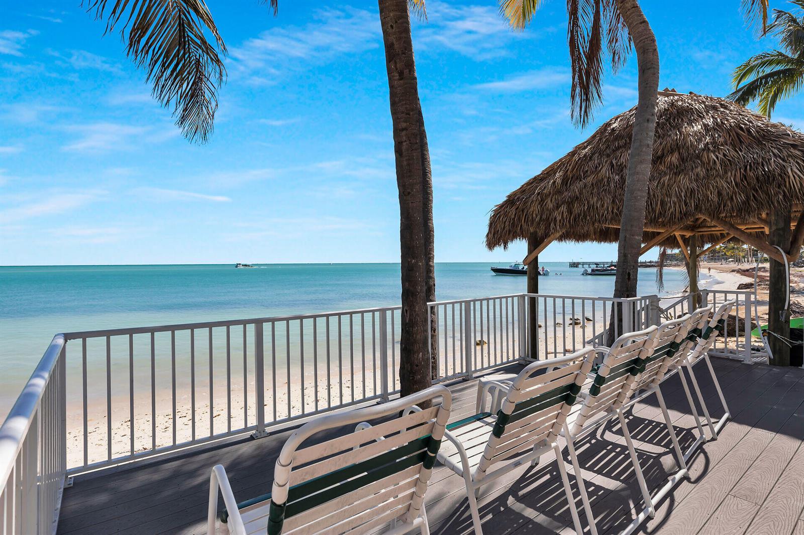 A wooden deck with lounge chairs overlooks a sandy beach, ocean, and a thatched-roof structure — 601 W Ocean Dr Drive # 203a, FL 33051, FL