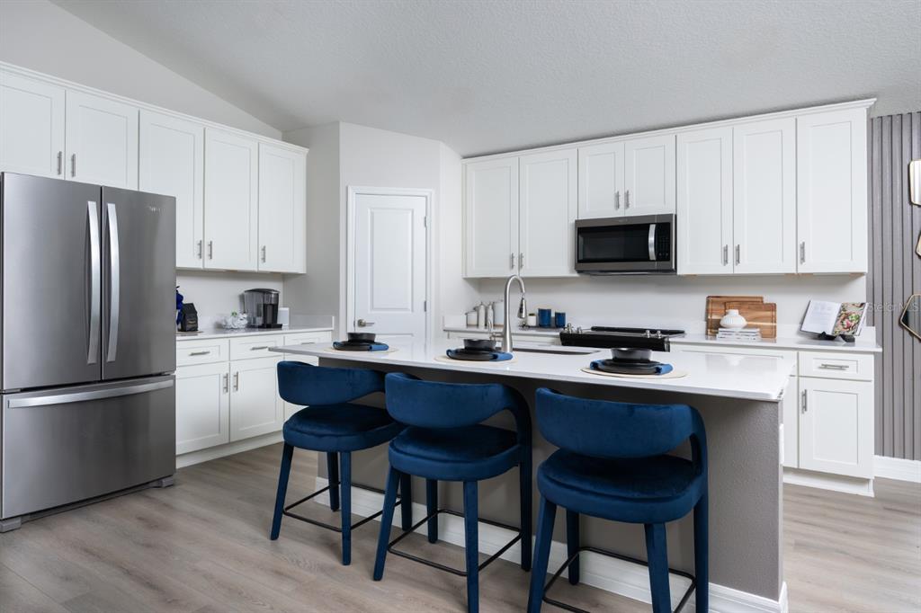 A modern kitchen features white cabinetry, stainless steel appliances, a quartz island, and blue stools — 6075 SW 97TH PLACE, OCALA, FL