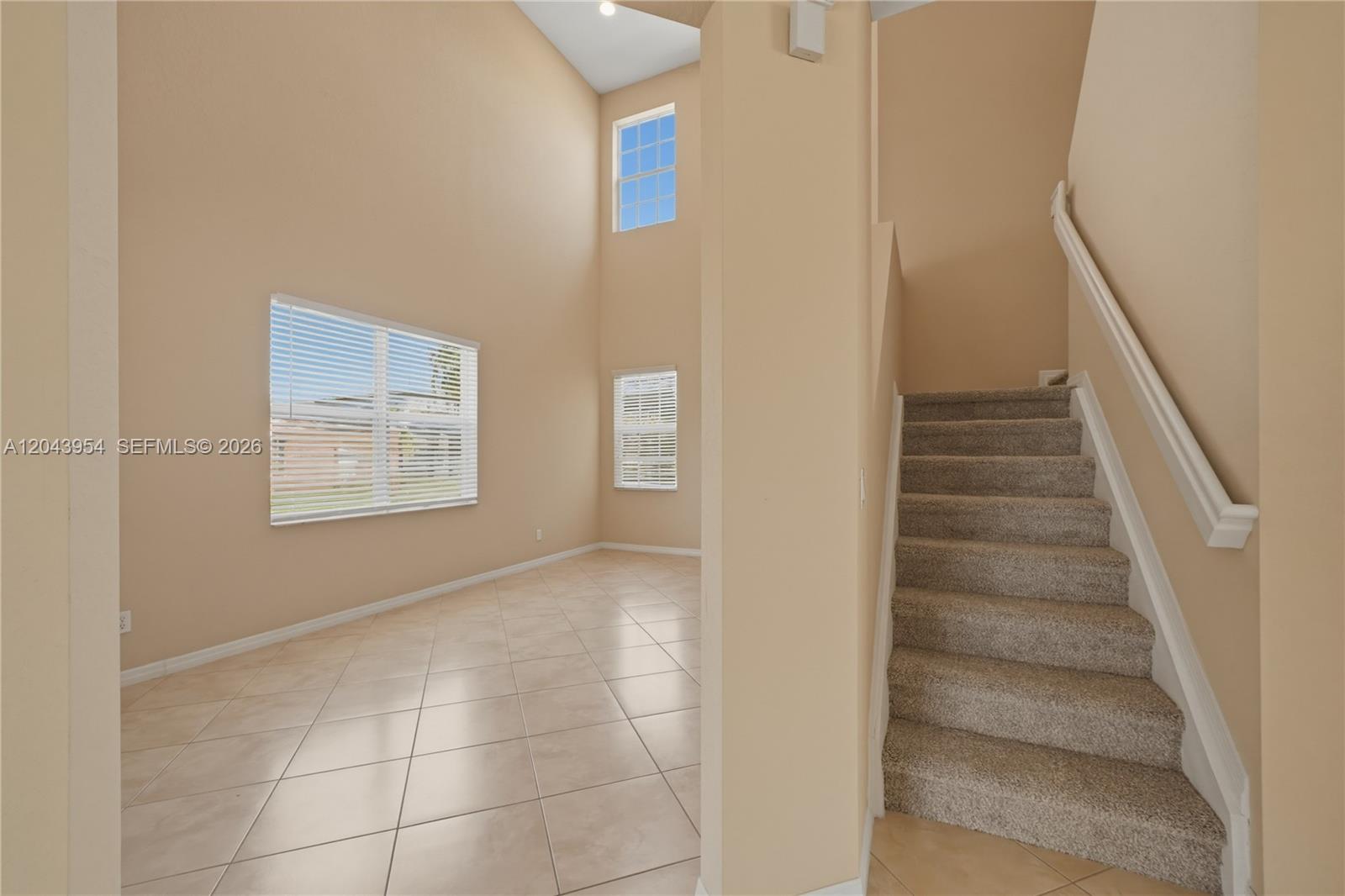 A room with beige tile flooring, high ceilings, and a carpeted staircase with a railing — 9317 Natures Way, Fort Pierce FL 34945, FORT PIERCE, FL