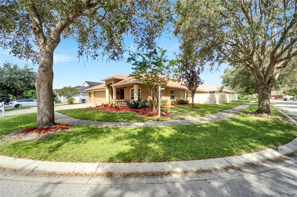 A tan single-story house with a two-car garage sits on a corner lot with trees — 12739 STANDBRIDGE DRIVE, RIVERVIEW, FL