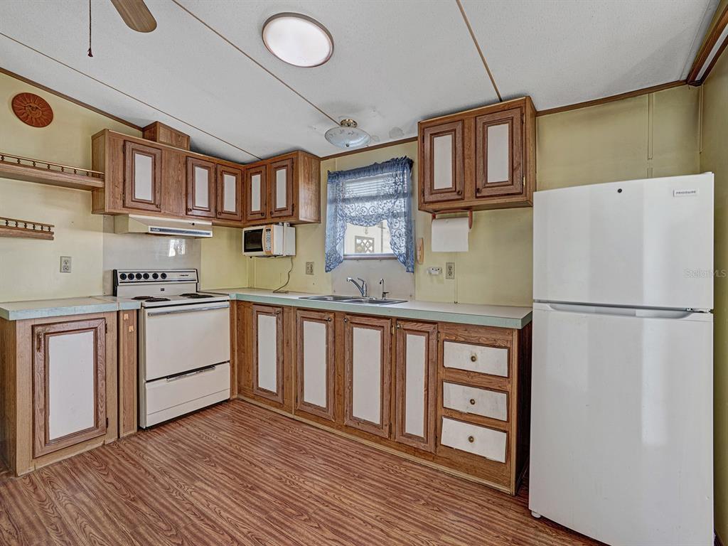 A kitchen features wood cabinets, white appliances, a window with blue curtains, and wood-look flooring — 10339 MARINETTE AVENUE, HUDSON, FL