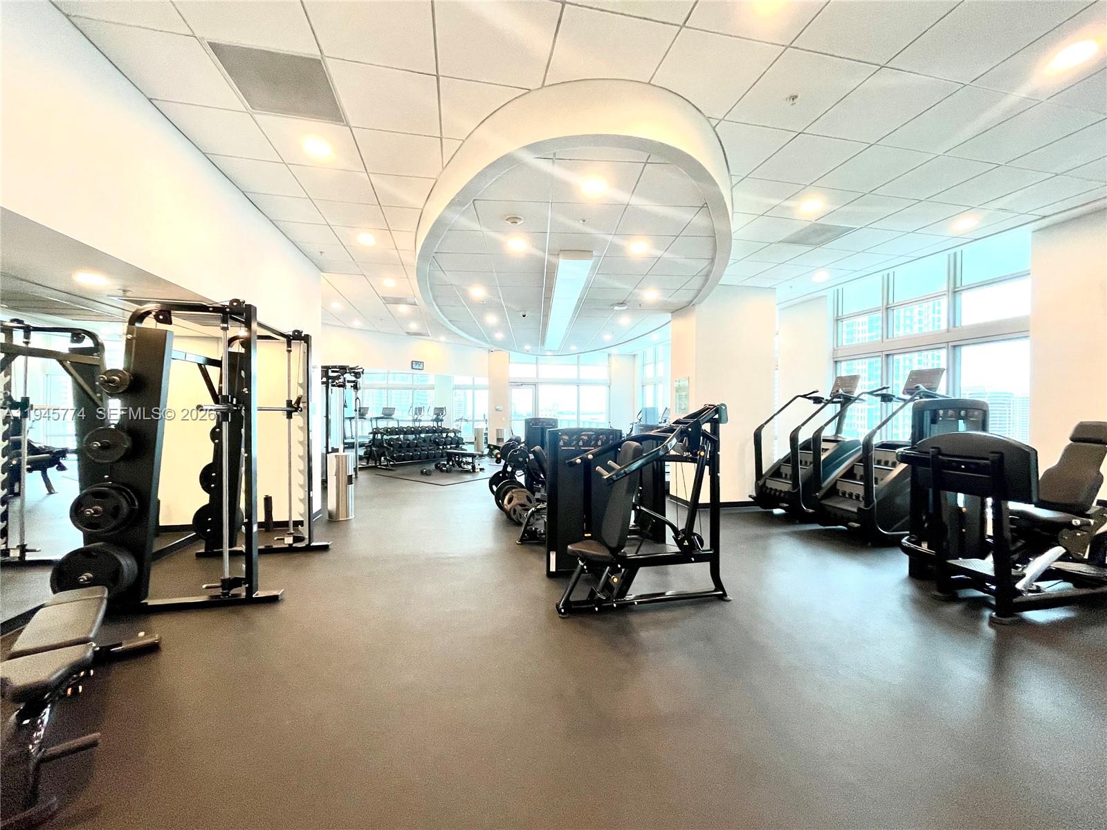 A modern fitness center features various strength training machines, free weights, and large windows — 300 S Biscayne Blvd # 1803, Miami FL 33131, MIAMI, FL