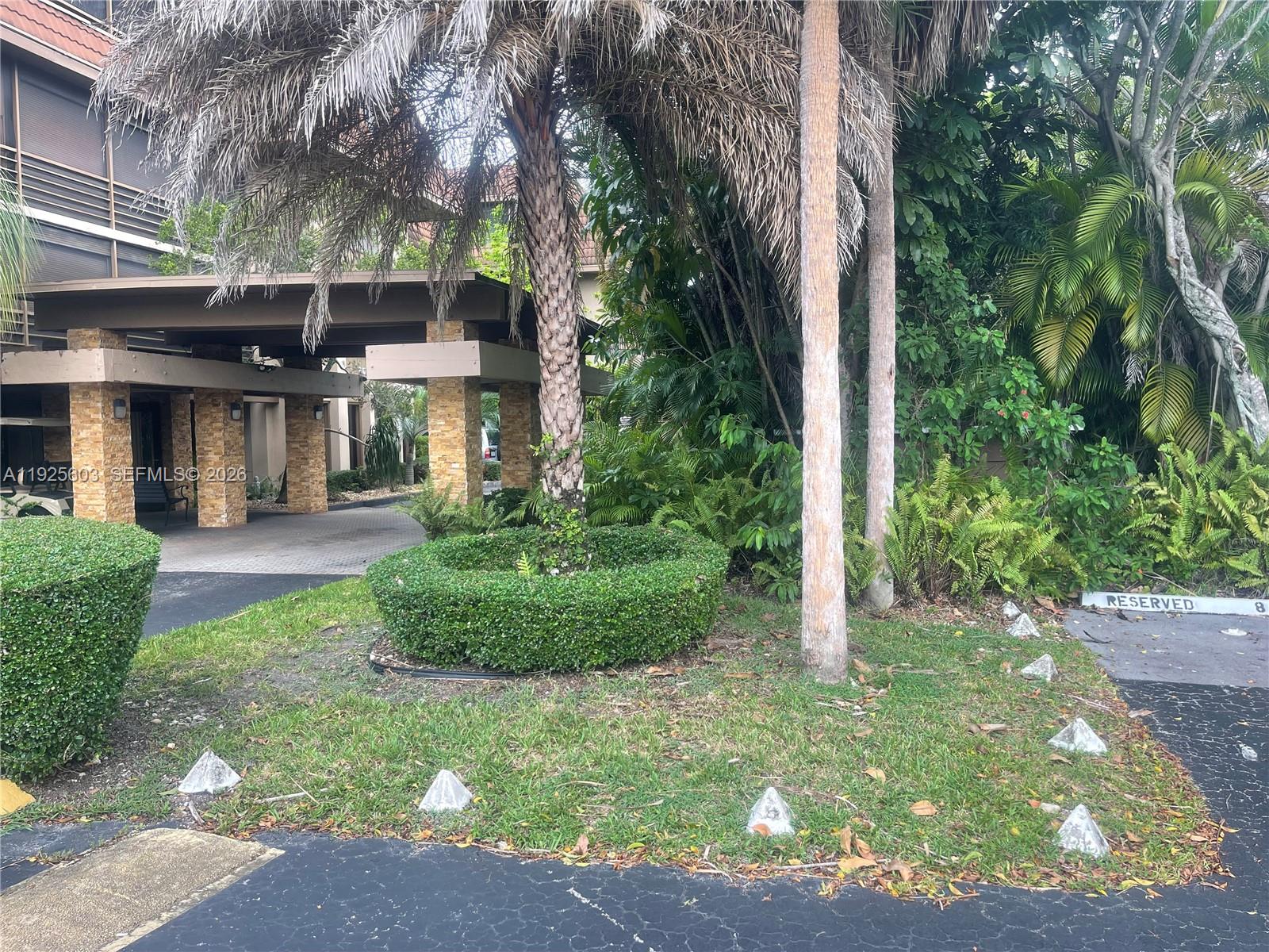 A covered entrance with stone pillars stands beside a palm tree and landscaped lawn — 661 NE 195th St # 309, Miami FL 33179, MIAMI, FL