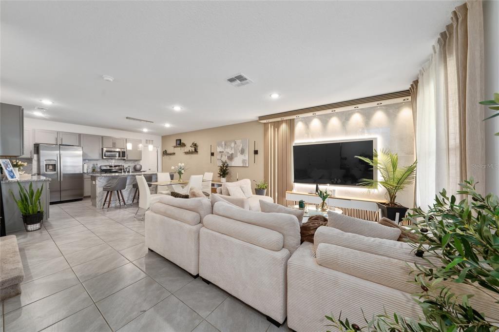 An open-concept living room and kitchen feature light tile flooring and a modern media wall — 15140 WINDFLOWER ALLEY, WINTER GARDEN, FL