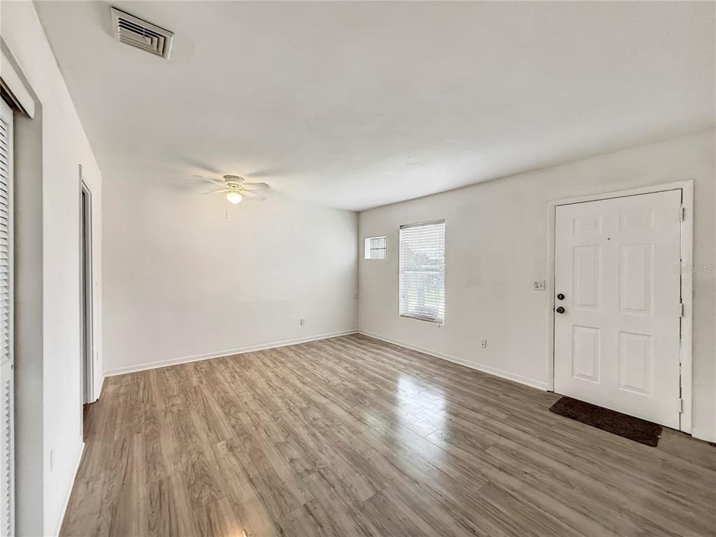 An empty living room features light-colored wood-look flooring, white walls, a ceiling fan, and a door — 889 W VILLAGE DRIVE, DELTONA, FL