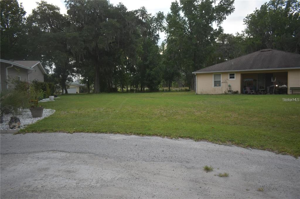 A grassy backyard sits between two houses with a sandy driveway in the foreground — LOT 25 BLK D STRATFORD COURT, WILDWOOD, FL