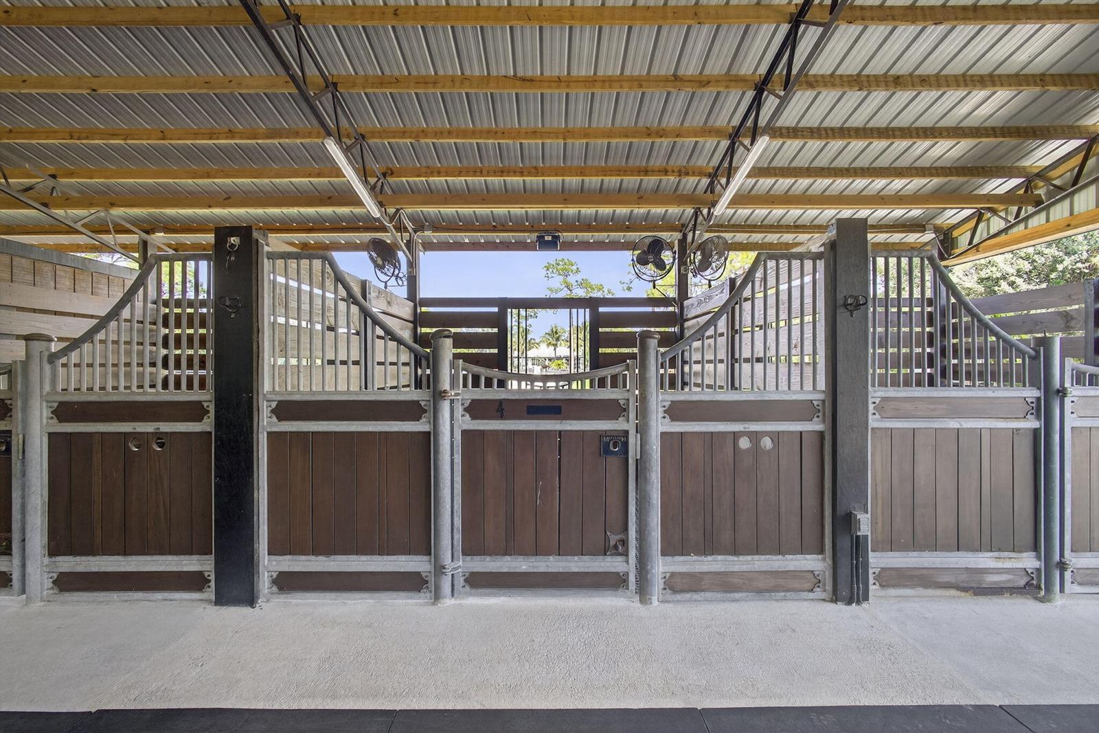 A covered horse stable features wooden stalls with metal frames and ceiling-mounted fans — 14346 Tripp Road N, FL 33470, FL