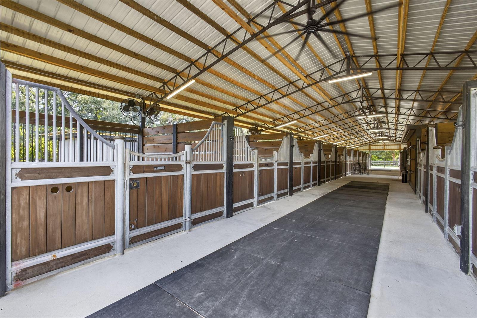 A long, covered horse stable aisle features wooden stalls with metal frames and rubber flooring — 14346 Tripp Road N, FL 33470, FL