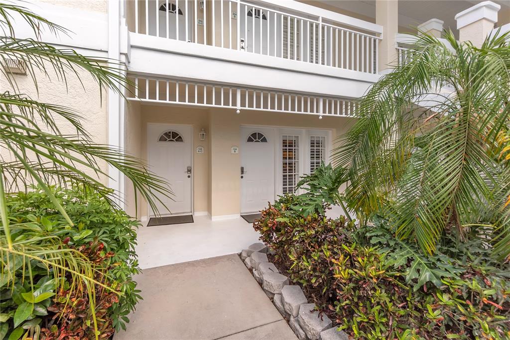 A ground-level exterior view of two white entry doors with a walkway and tropical landscaping — 3401 54TH DRIVE W UNIT 104, BRADENTON, FL