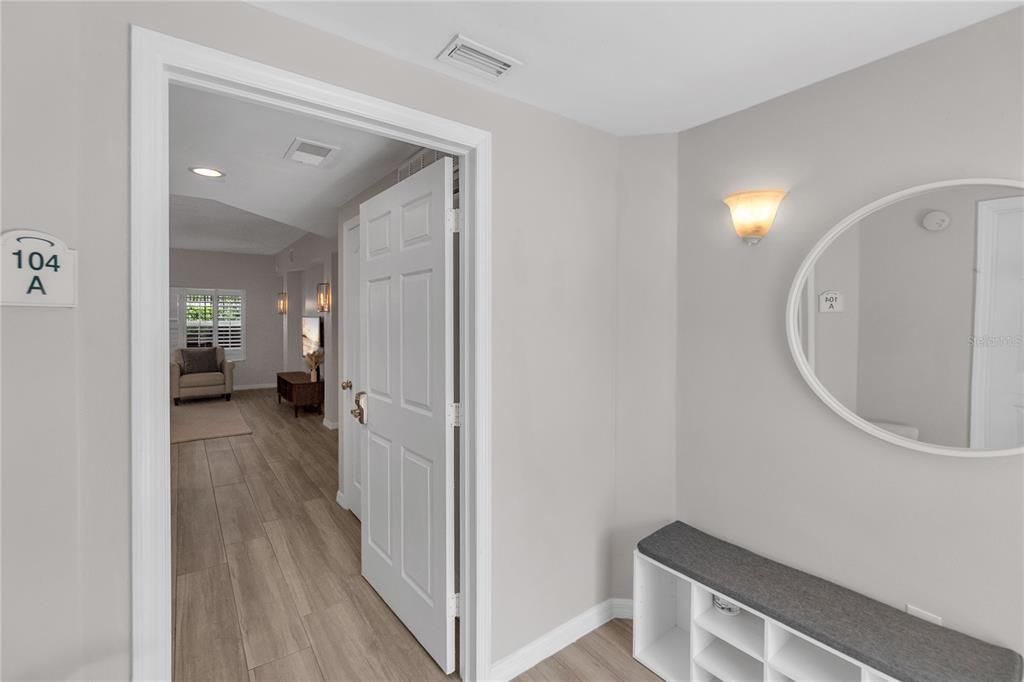 An entryway features a white door, a bench with storage, and a round wall mirror — 3401 54TH DRIVE W UNIT 104, BRADENTON, FL