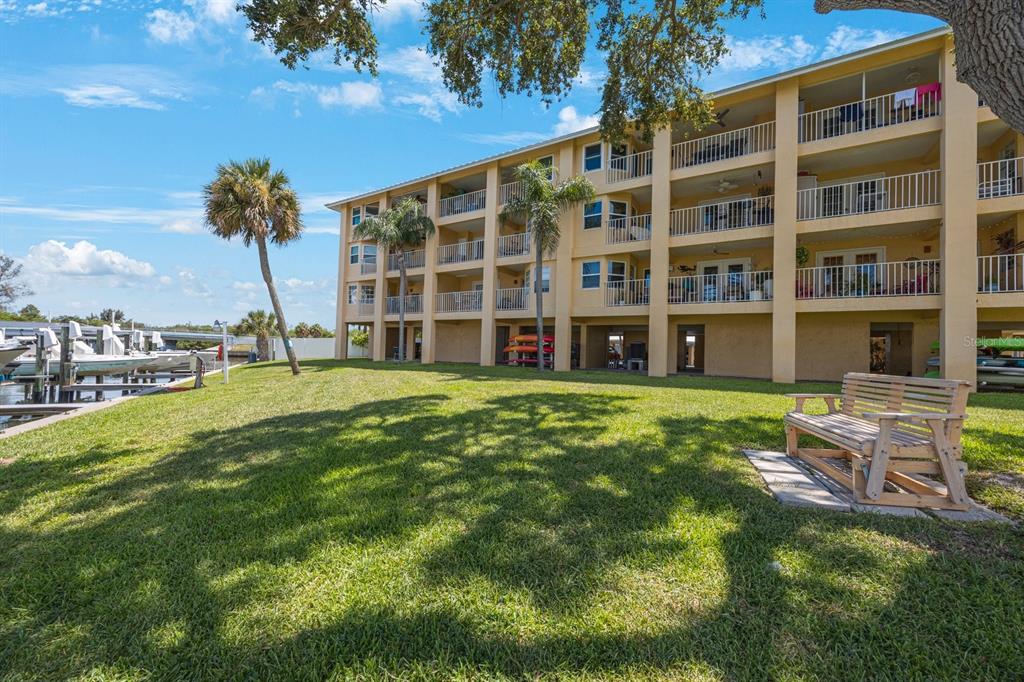 A multi-story apartment building with balconies overlooks a grassy lawn, a wooden bench, and boats — 1125 PINELLAS BAYWAY S UNIT 305, ST PETERSBURG, FL