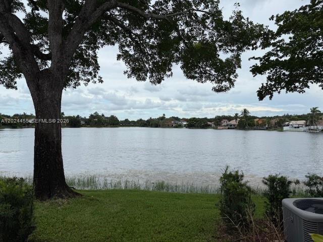 A large tree stands on a grassy lawn overlooking a calm lake with distant houses — 2856 Cambridge Ln, Cooper City FL 33026, COOPER CITY, FL