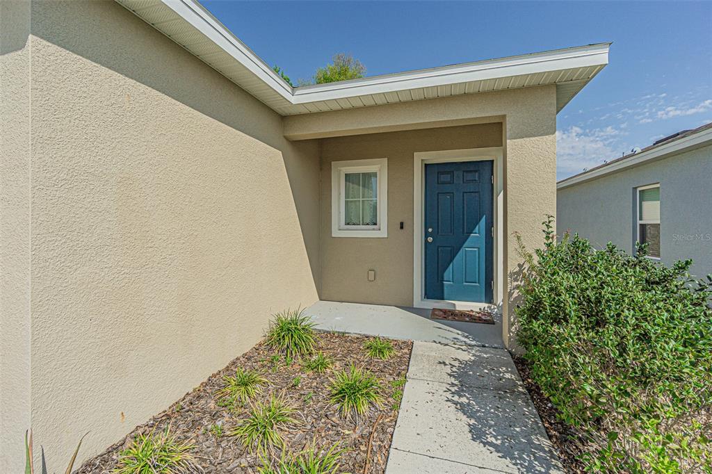 A concrete walkway leads to the blue front door of a tan stucco house — 35045 JOMAR AVENUE, ZEPHYRHILLS, FL