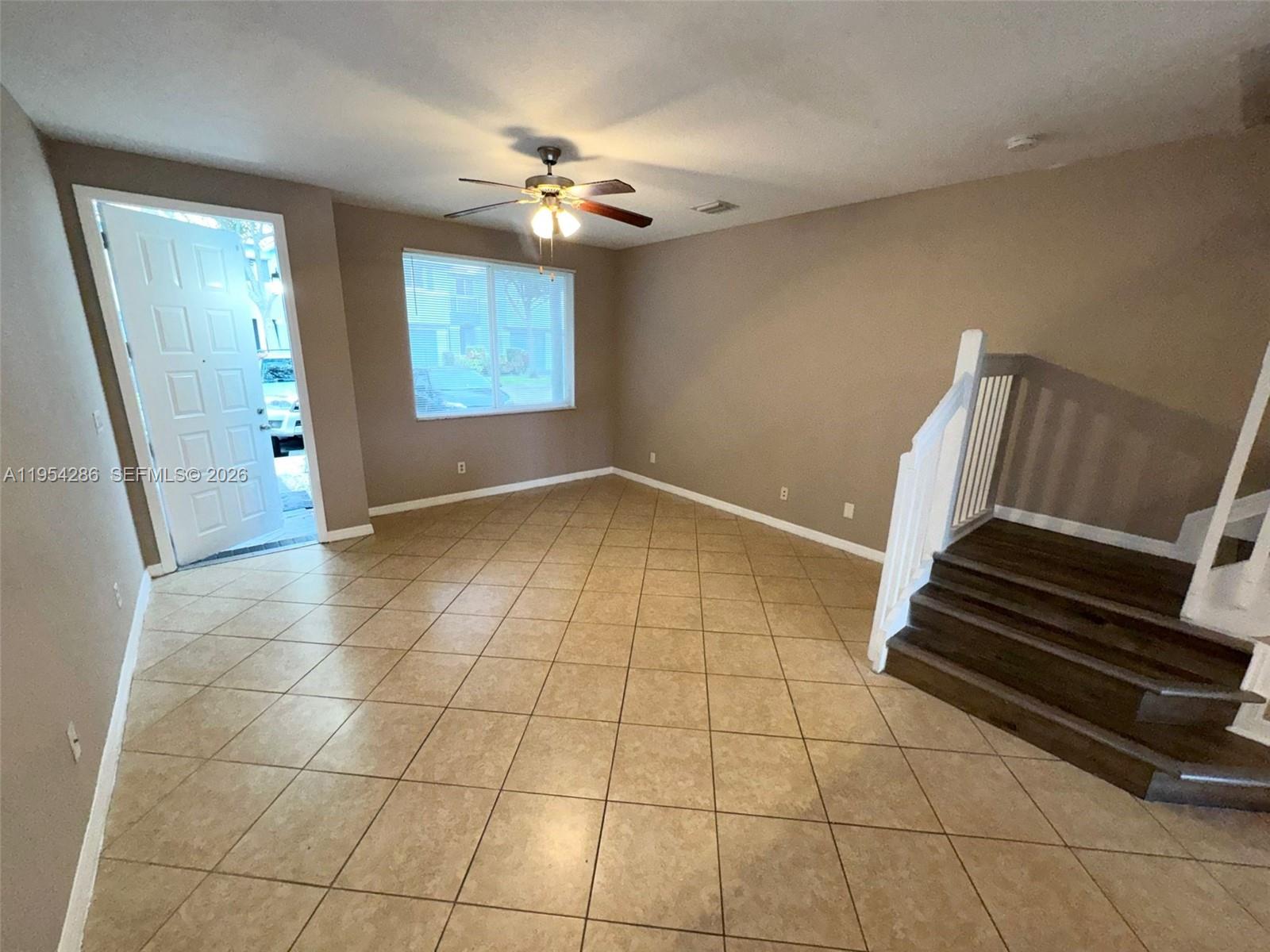 An empty living room features tan tile flooring, beige walls, a ceiling fan, and stairs — 3622 Oleander Ter, Riviera Beach FL 33404, RIVIERA BEACH, FL