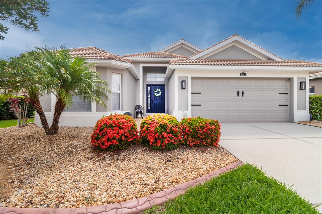 A single-story house exterior features a gray garage, blue front door, and red flowering shrubs — 8964 STONE HARBOUR LOOP, BRADENTON, FL