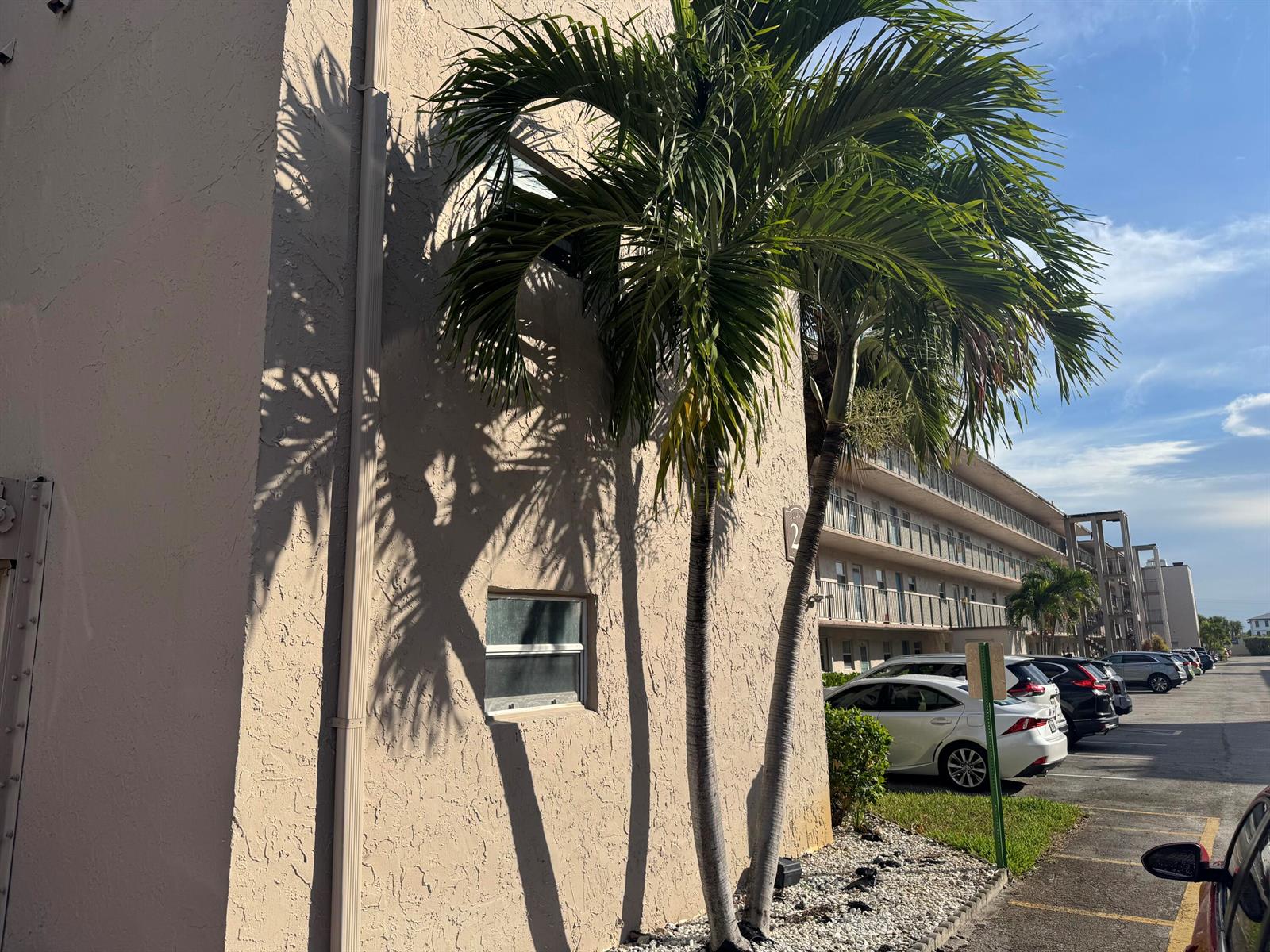 Exterior view of a beige apartment building with palm trees and a parking lot — 725 Lori Drive # 107, Palm Springs FL 33461, PALM SPRINGS, FL