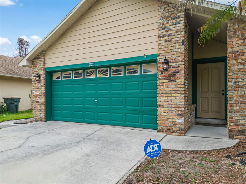 A house exterior features a green garage door, brick accents, and a beige front door — 1321 WOOD LAKE CIRCLE, ST CLOUD, FL