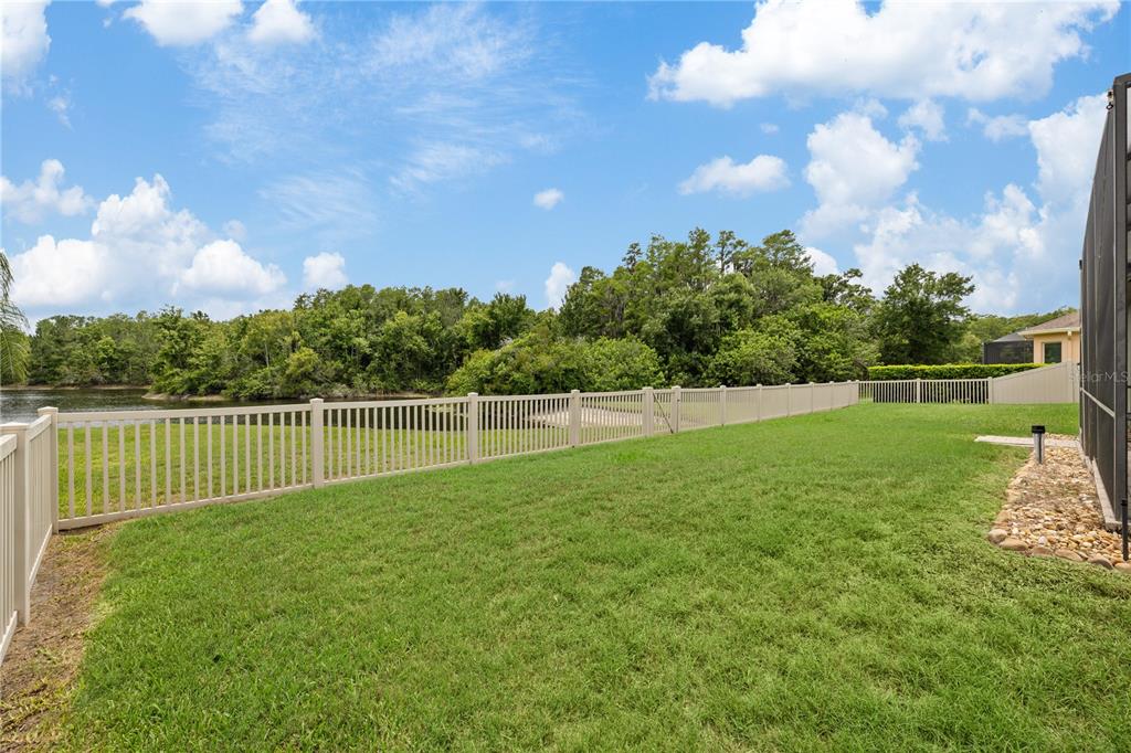 A grassy backyard features a vinyl fence bordering a pond and dense trees — 3015 LYNWOOD COURT, LAND O LAKES, FL