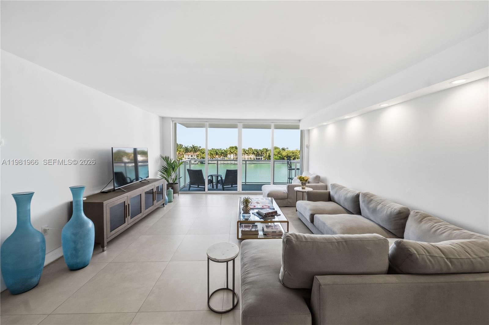 A living room features a grey sectional sofa, a media console, and a waterfront balcony — 5750 Collins Ave # 4F, Miami Beach FL 33140, MIAMI BEACH, FL