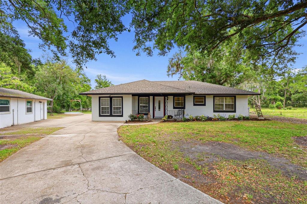 A single-story white house with a dark roof and driveway sits under large oak trees — 30151 LYNNE DRIVE, WESLEY CHAPEL, FL