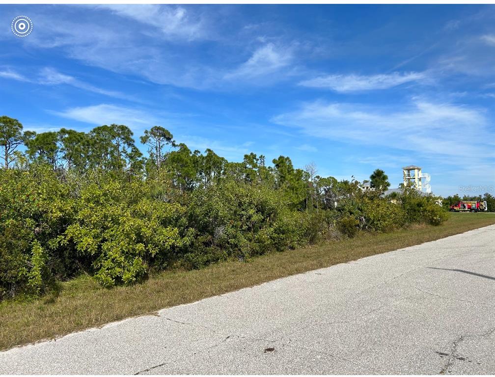 A paved road runs alongside a vacant, overgrown lot with trees under a blue sky — 16268 RENNIE ROAD, PORT CHARLOTTE, FL