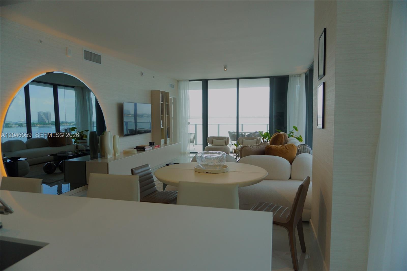 A modern living and dining area features a circular mirror, white furniture, and floor-to-ceiling windows — 725 NE 24 St # 401, Miami FL 33137, MIAMI, FL