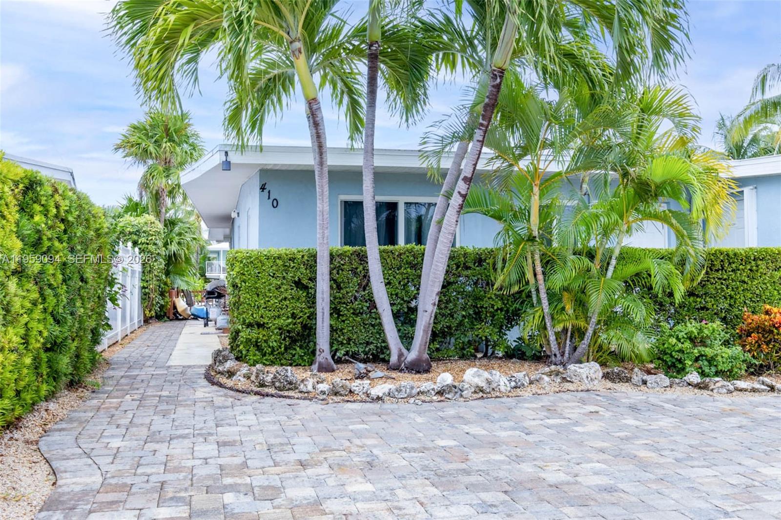 A light blue house exterior features a paved driveway, palm trees, and a trimmed hedge — 410 10th St, Key Colony FL 33051, KEY COLONY, FL