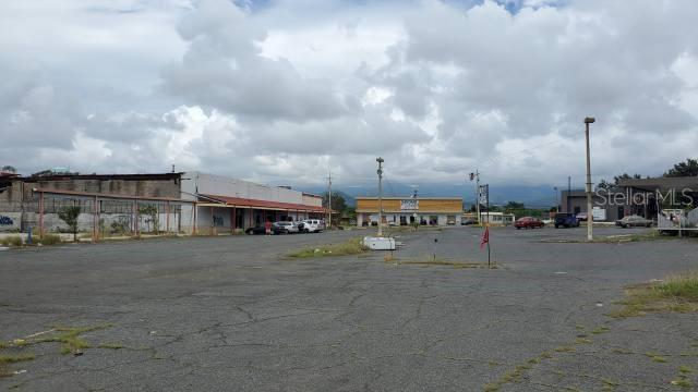 A large, paved parking lot sits in front of several commercial buildings under a cloudy sky — PR-116 INT. PR-121, YAUCO, PR