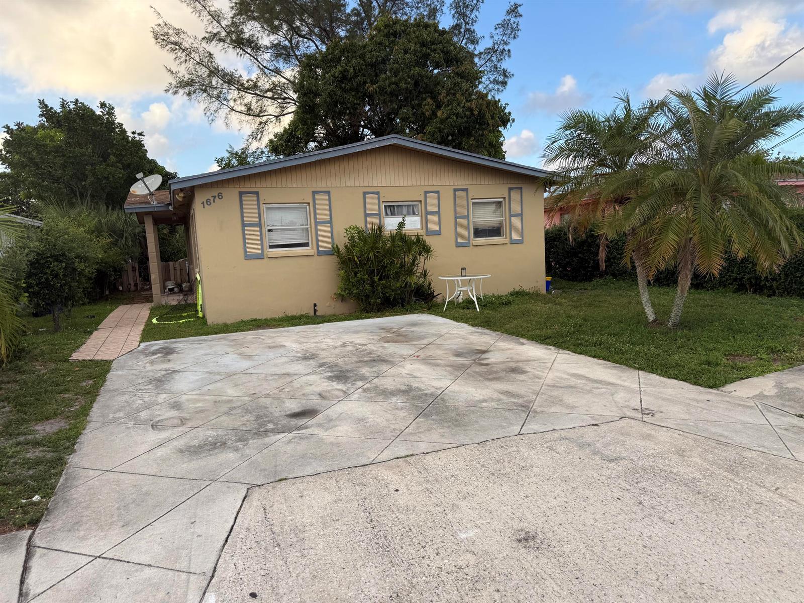 A tan, single-story house with blue shutters and a concrete driveway sits under a blue sky — 1676 W 10th Street, Riviera Beach FL 33404, RIVIERA BEACH, FL