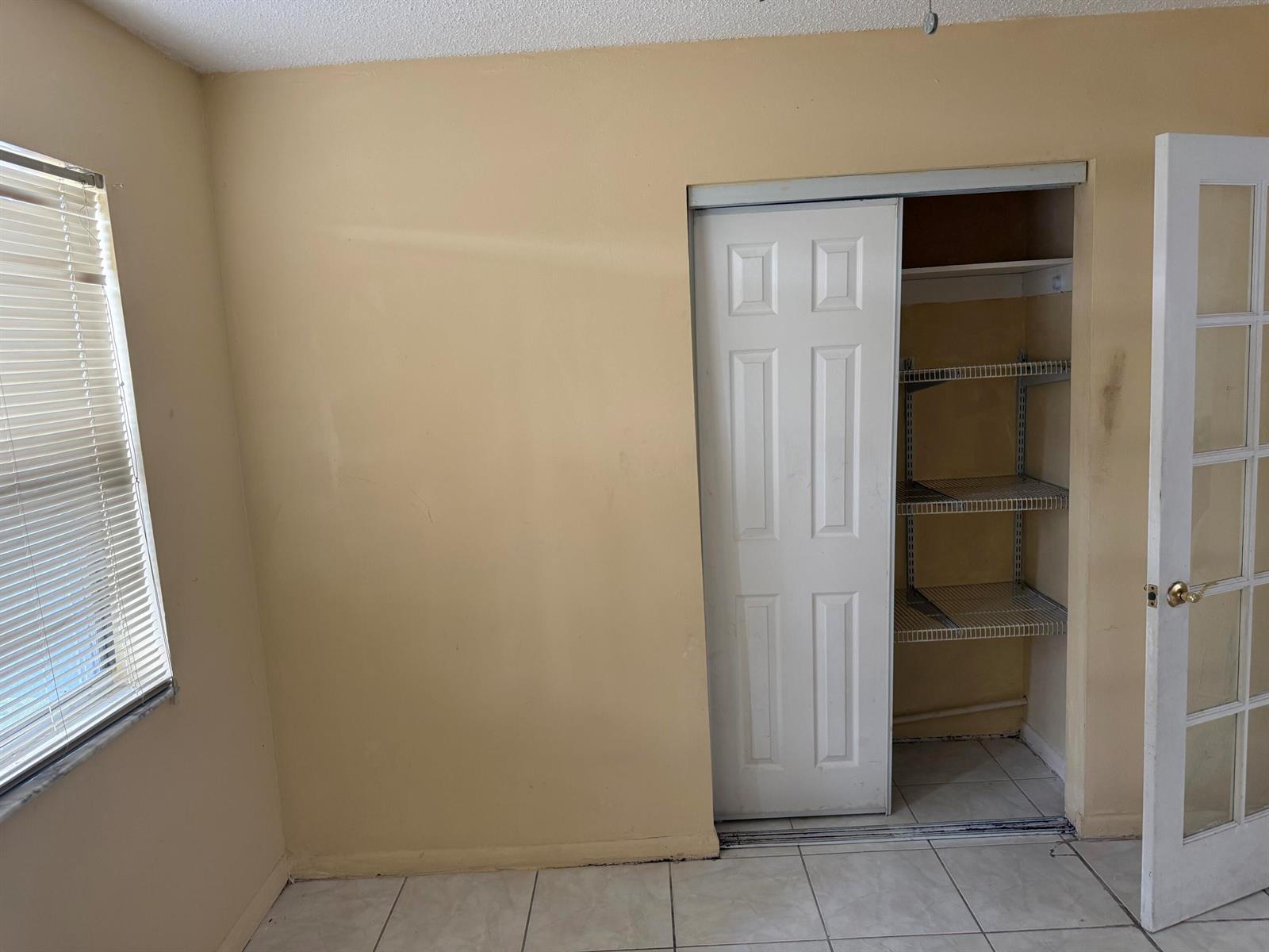 A room with beige walls, tiled flooring, a window with blinds, and an open closet — 1676 W 10th Street, Riviera Beach FL 33404, RIVIERA BEACH, FL