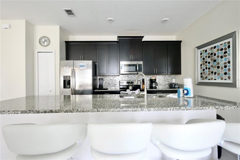 A kitchen features dark cabinets, granite countertops, stainless steel appliances, and three white bar stools — 9063 DOGLEG DRIVE, DAVENPORT, FL