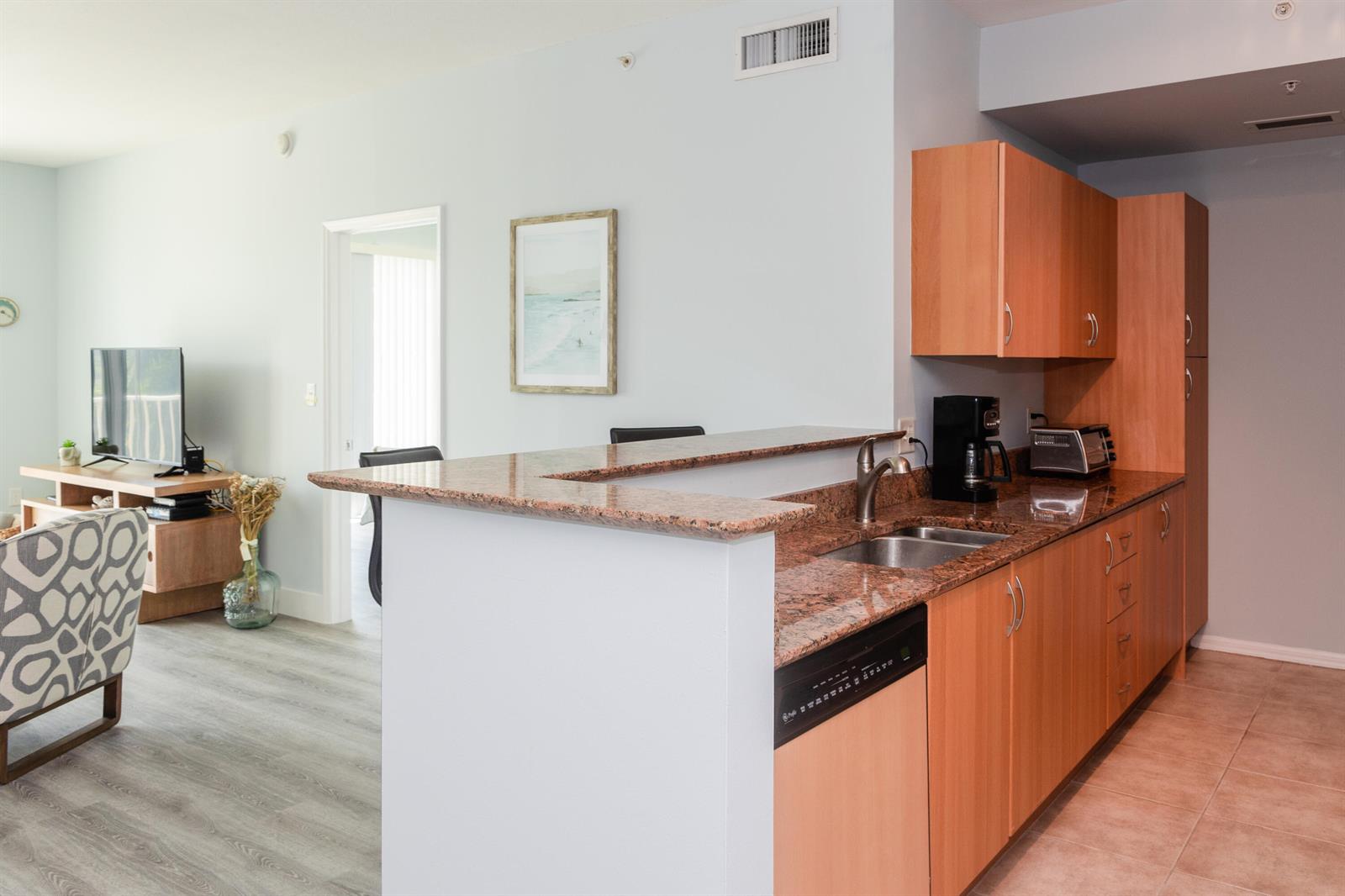 A kitchen with granite countertops and wood cabinets overlooks a living area with a television — 802 W Windward Way # 311, Lantana FL 33462, LANTANA, FL