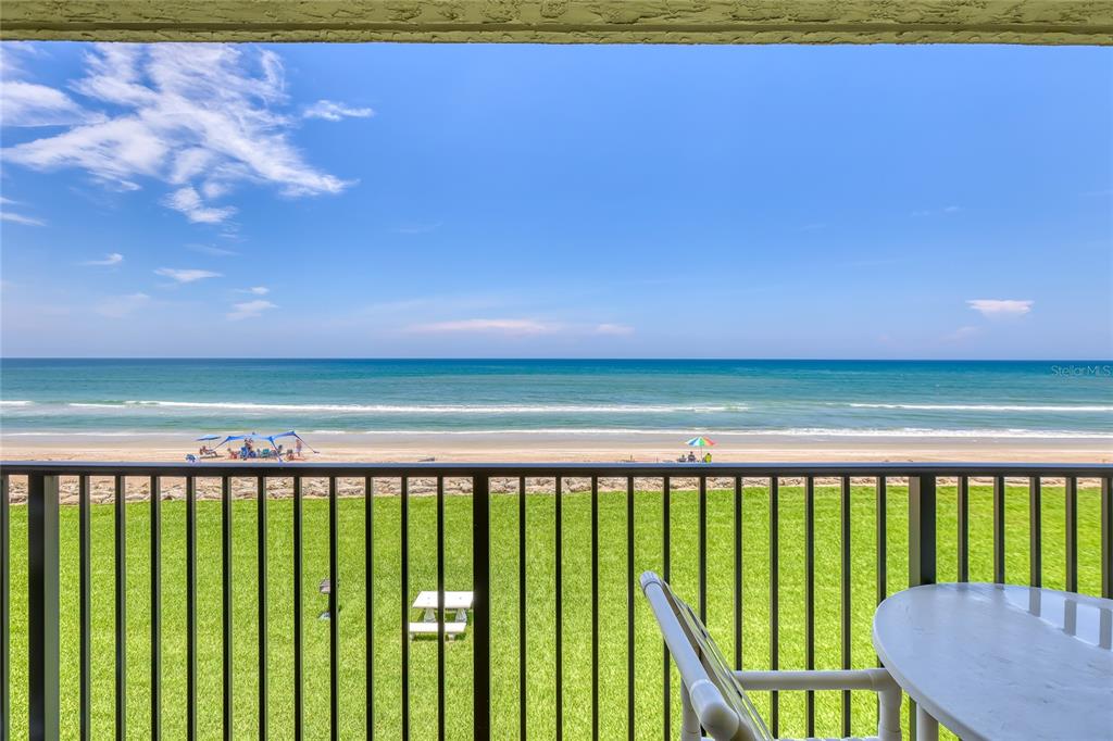 A balcony with a table and chairs overlooks a grassy lawn and the ocean beach — 6727 TURTLEMOUND ROAD UNIT 318, NEW SMYRNA BEACH, FL
