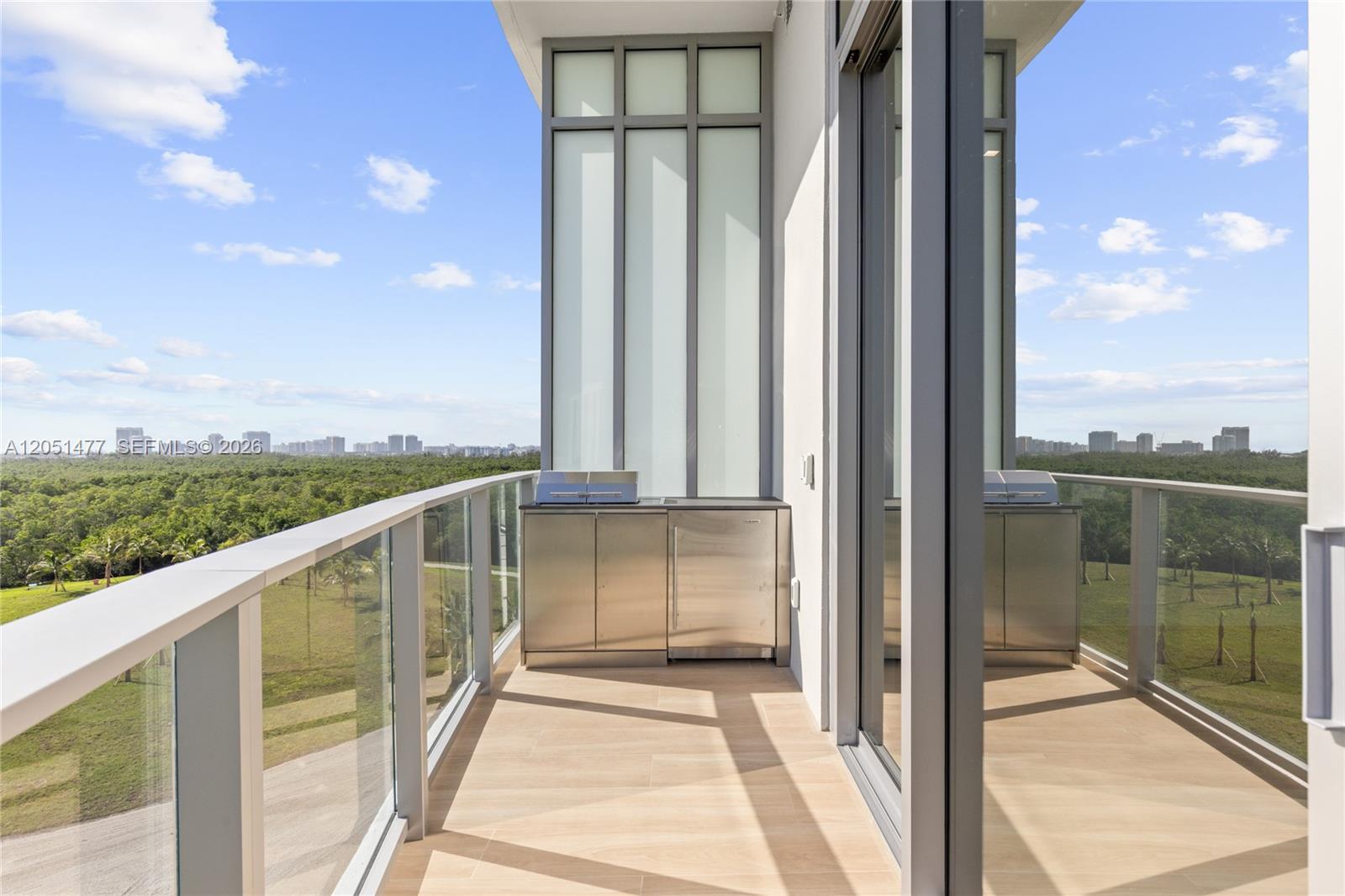 A balcony features a stainless steel outdoor kitchen, glass railings, and views of greenery — 2411 Laguna Cr # 611, Miami FL 33181, MIAMI, FL