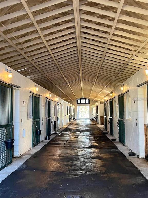 A long, covered horse stable aisle features concrete flooring and rows of green stall doors — 5540 SE 157TH STREET ROAD UNIT (Option 1), SUMMERFIELD, FL